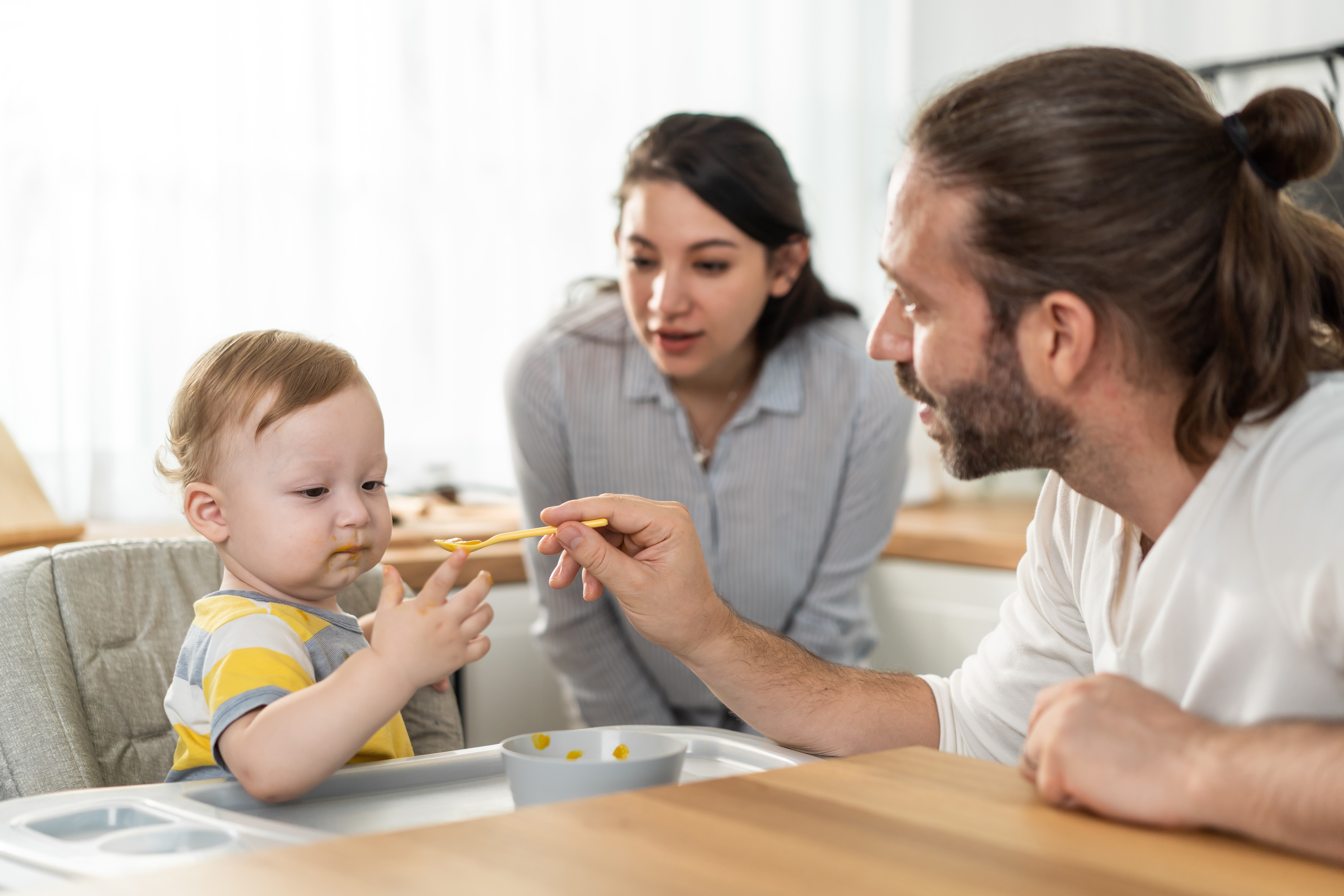 Caucasian young baby toddler eating healthy foods in kitchen at home. Little adorable cute kid boy infant sitting on children chair alone, enjoy having lunch in house. Child development skill concept.